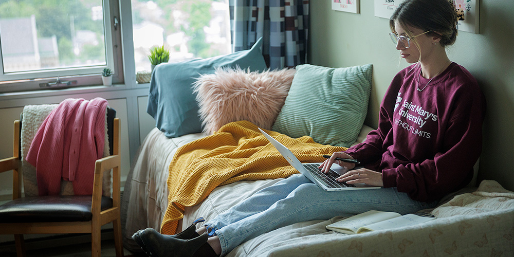 A student sits comfortably on a bed in residence, using a laptop.