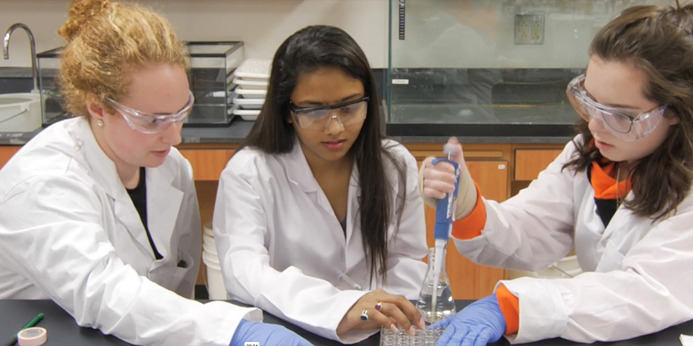 Three students in lab coats and safety glasses are filling vials with a pipette pump.