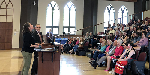 Two people stand at a podium in front of a seated audience.