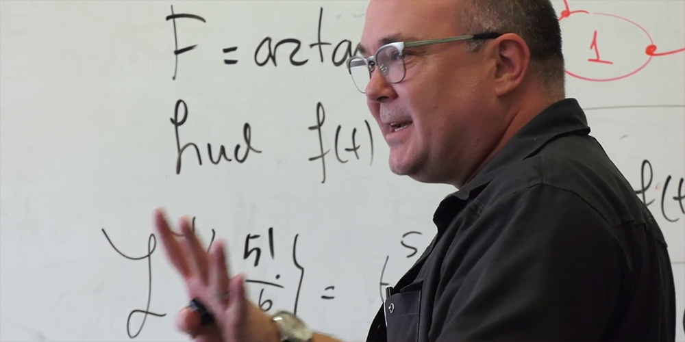 A professor stands in front of a white board with math equations.