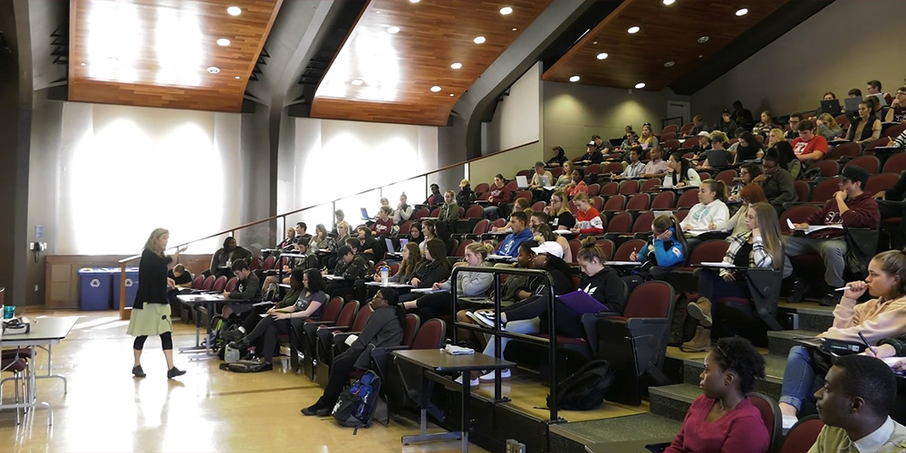 A professor lectures students in a large, light-filled classrrom.